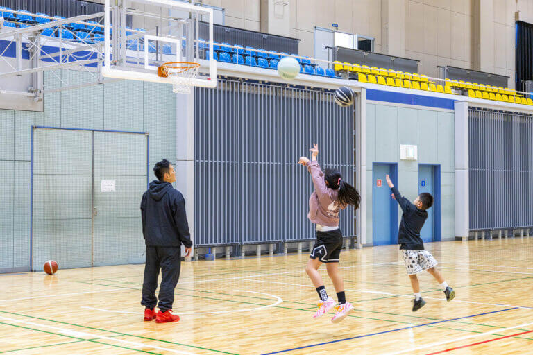 三豊市総合体育館アリーナでバスケットボールを楽しむ家族。バスケットゴールへボールを投げる女の子と男の子と、見守る父親